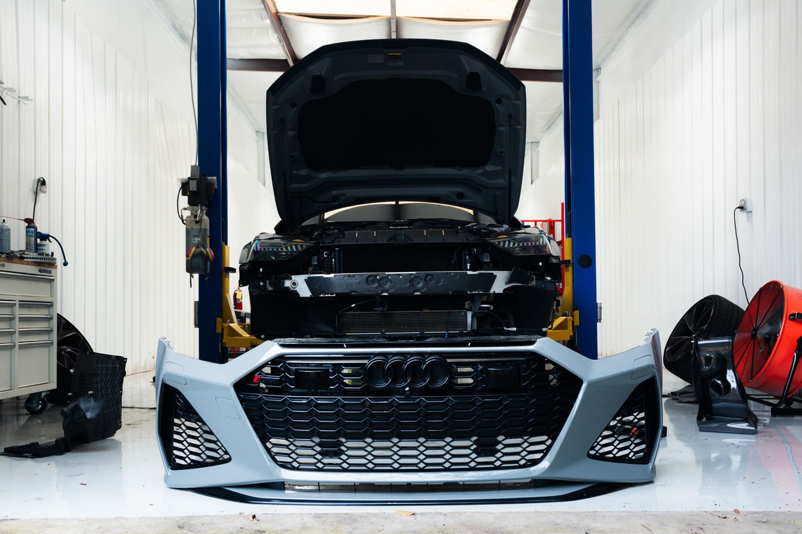 Grey Audi RS7 on a two-post lift with the hood open and the front bumper off, set on the floor in front of it
